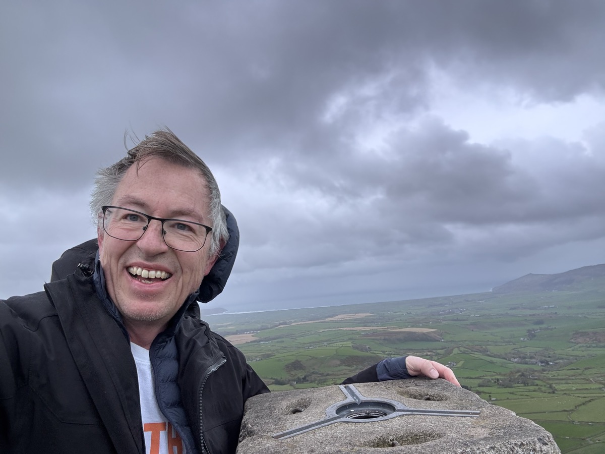 Julian at the trig point on the summit, Welsh coastline behind him