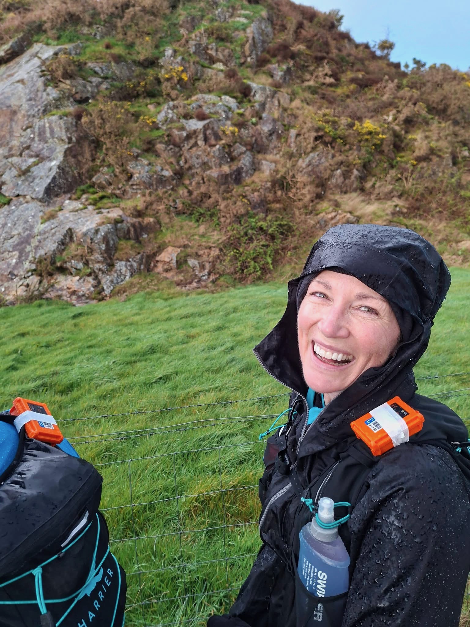 Cass beaming under a soaked black hood, Harrier vest and dibber clips on, Welsh rocky outcrop behind her